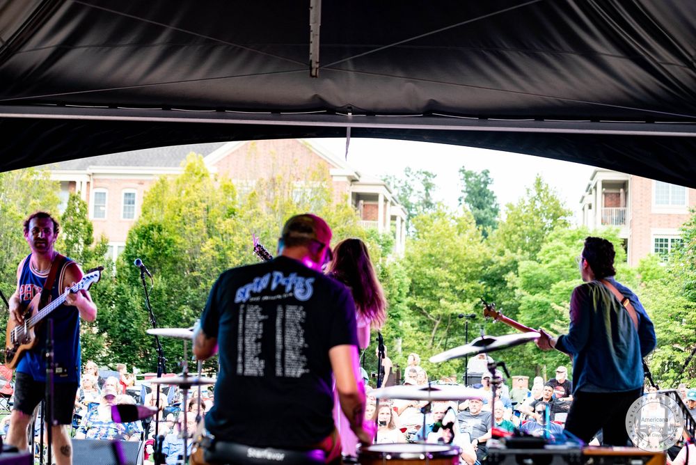 Band performs on outdoor stage before large crowd with trees and buildings in background.