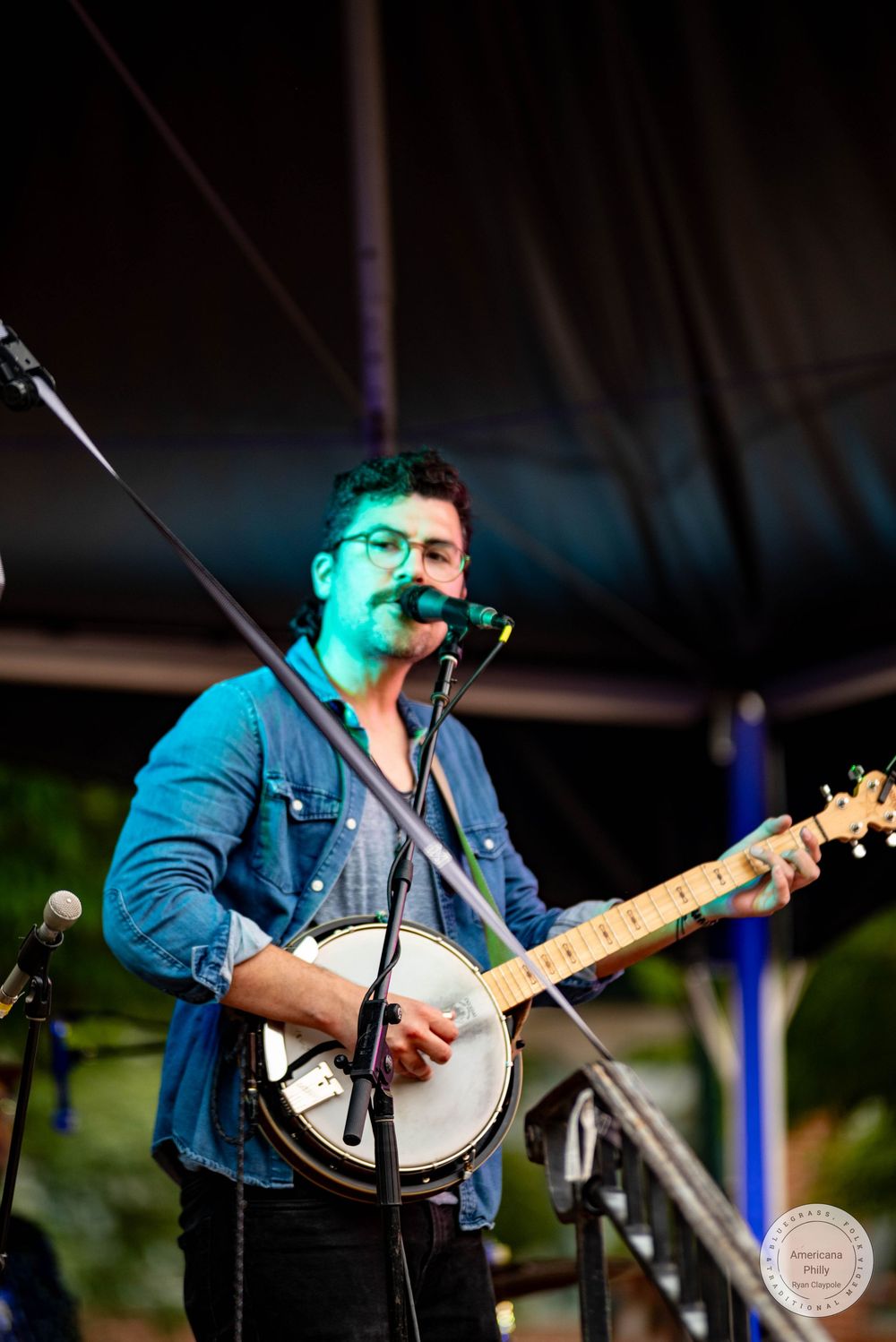 Man in blue denim shirt playing banjo and singing into microphone on outdoor stage at live music event.