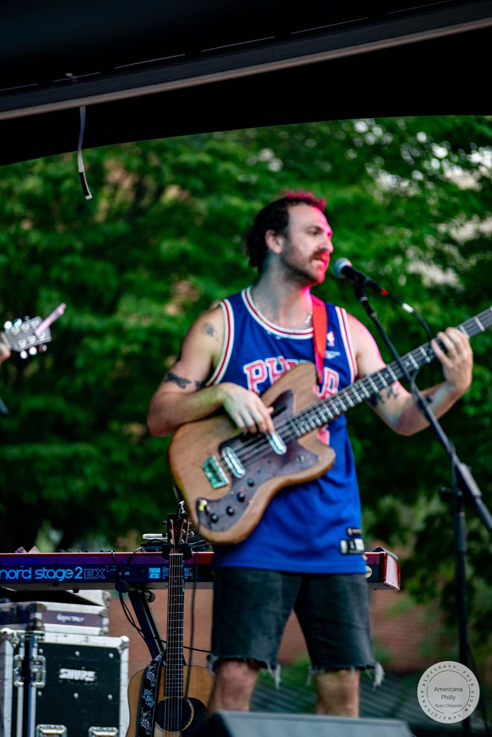 Bearded male guitarist performing on outdoor stage wearing blue basketball jersey and denim shorts with green trees in background.