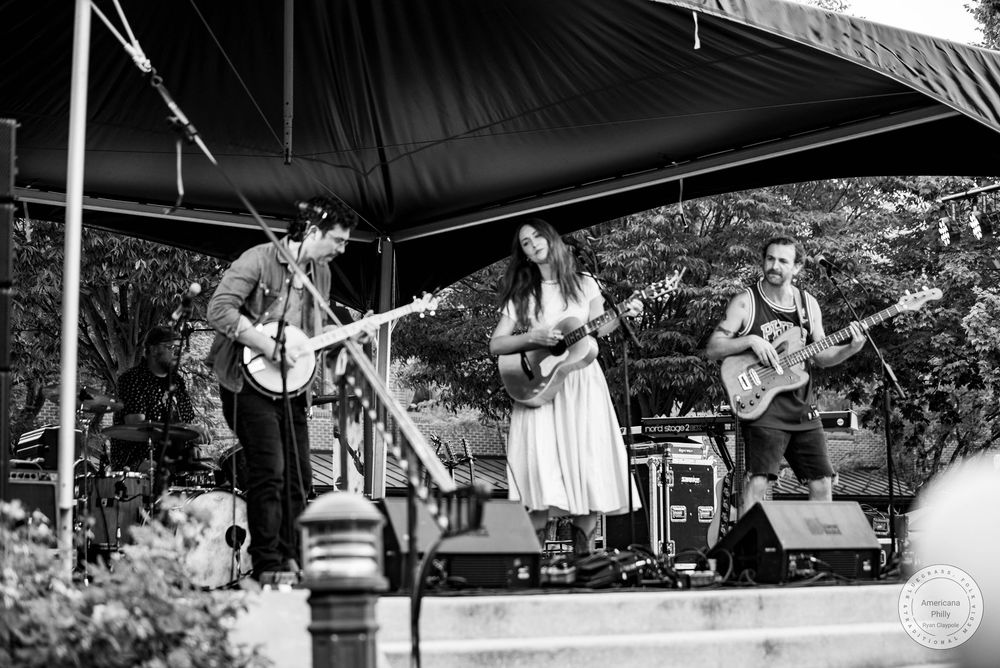 Black and white photo of a three-piece band performing on an outdoor stage, featuring two guitarists and a female vocalist.