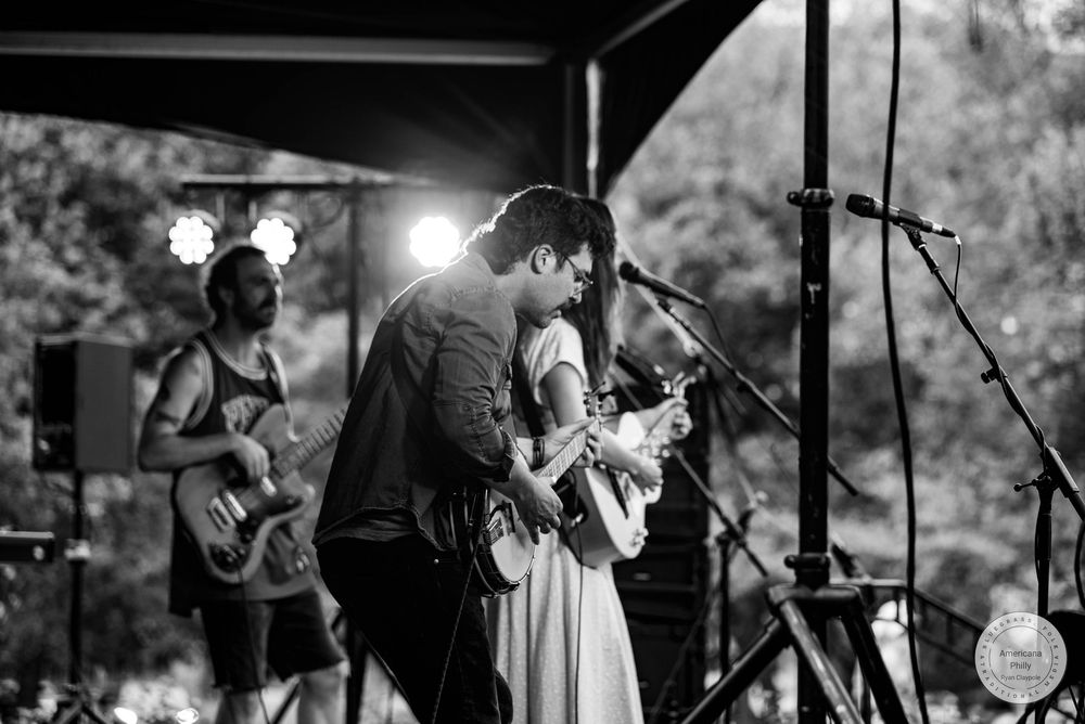 Black and white photo of musicians performing on stage, with guitarist and singer sharing a microphone at an outdoor concert.