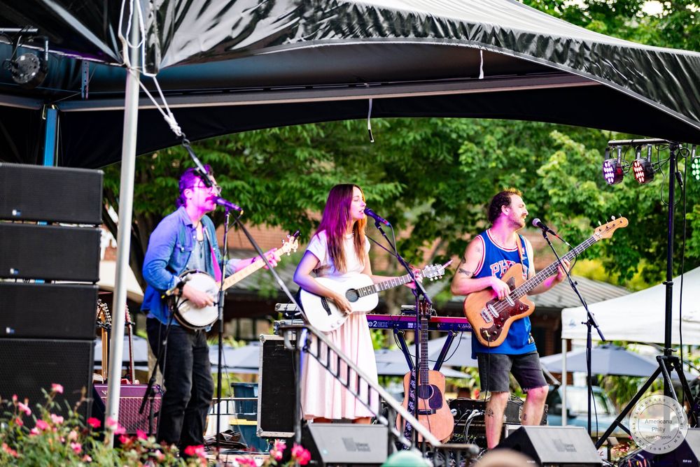 Three musicians perform on an outdoor stage, playing banjo, guitar, and keyboard at a sunny live music festival.