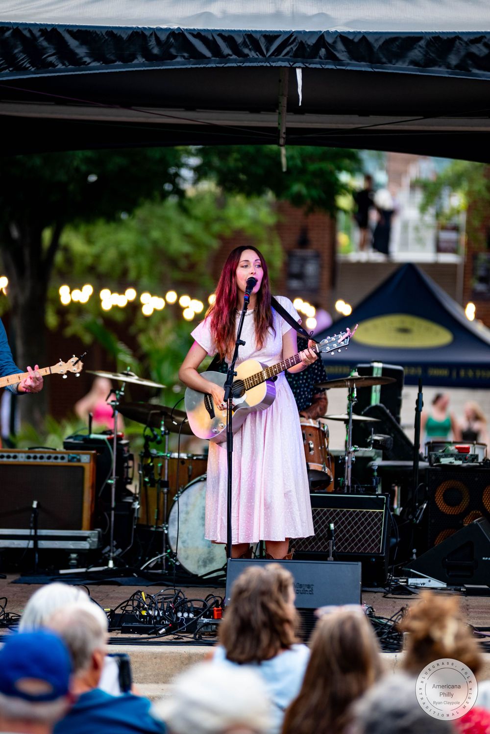 Female singer with red hair performs on outdoor stage in pink dress, playing acoustic guitar at a live music event.