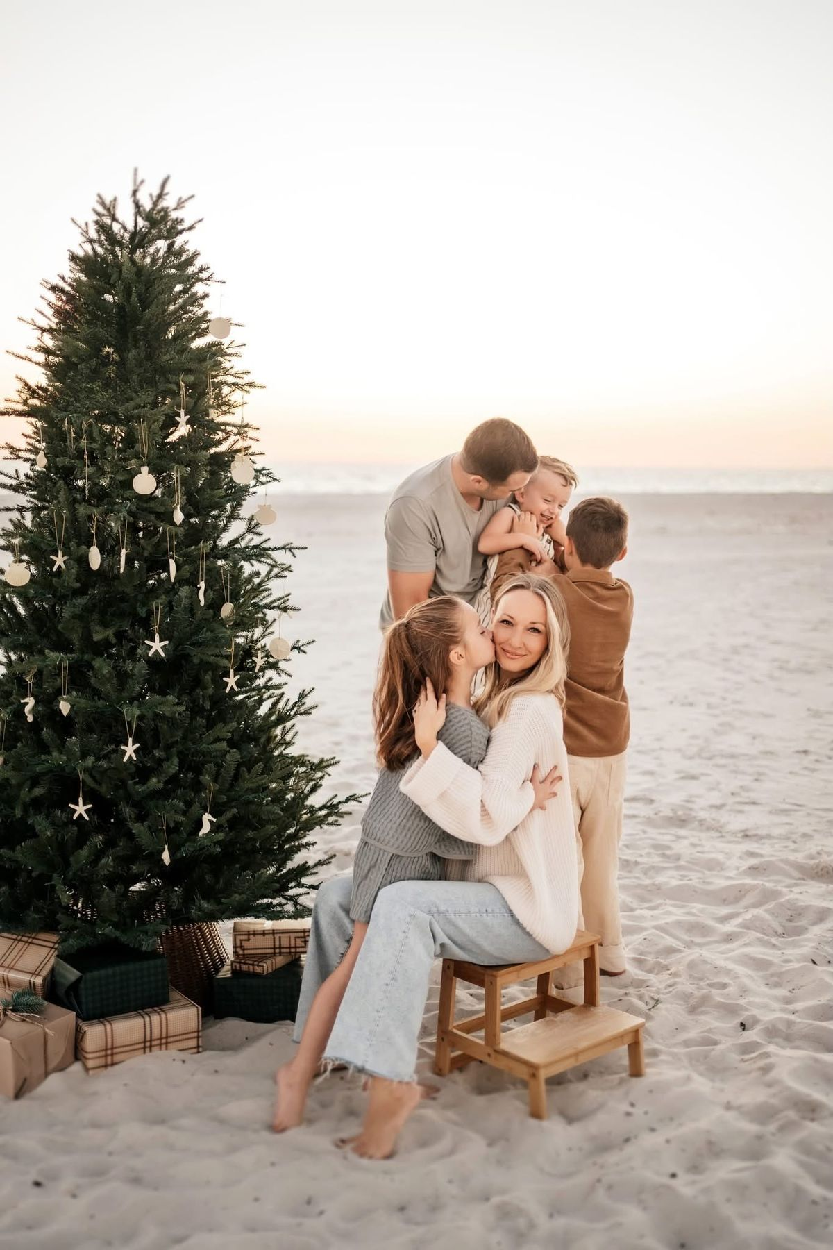 Family of four reads a book together on a white rug beside a decorated Christmas tree with gifts.