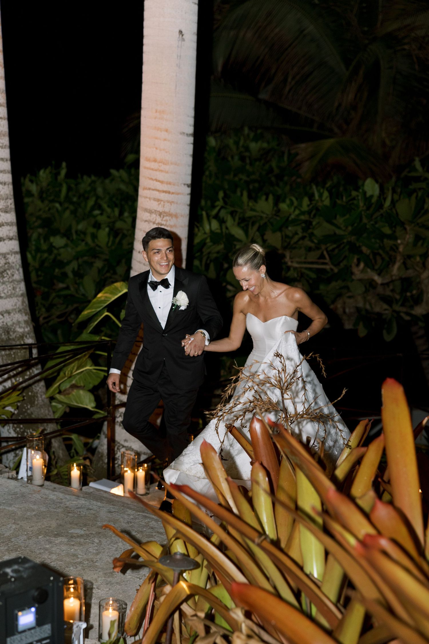 Bride in backless lace gown and groom in white shirt walk up candlelit stairs at night wedding reception.