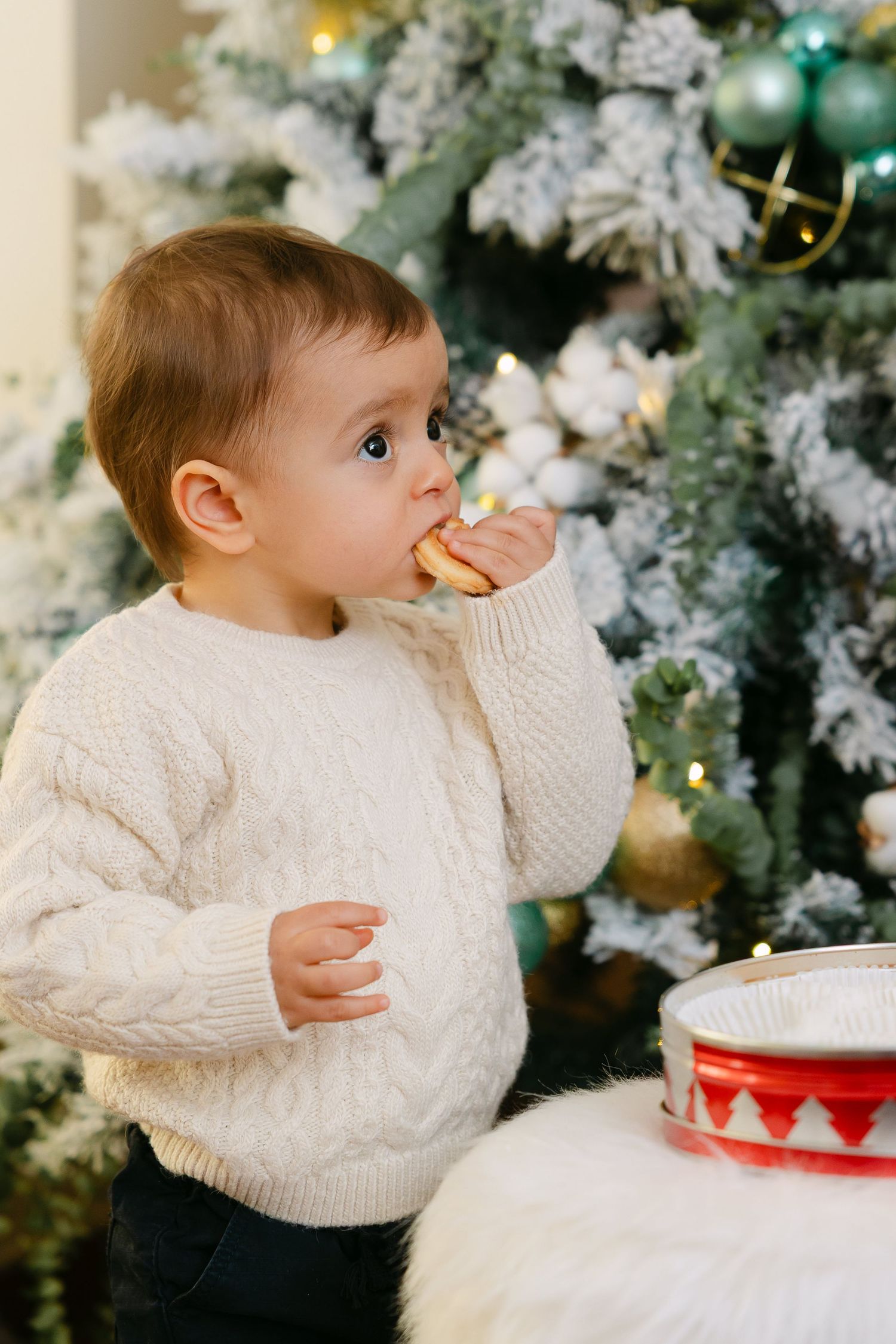 Two young siblings in white knit sweaters sharing cookies from a festive tin while sitting together in an armchair.