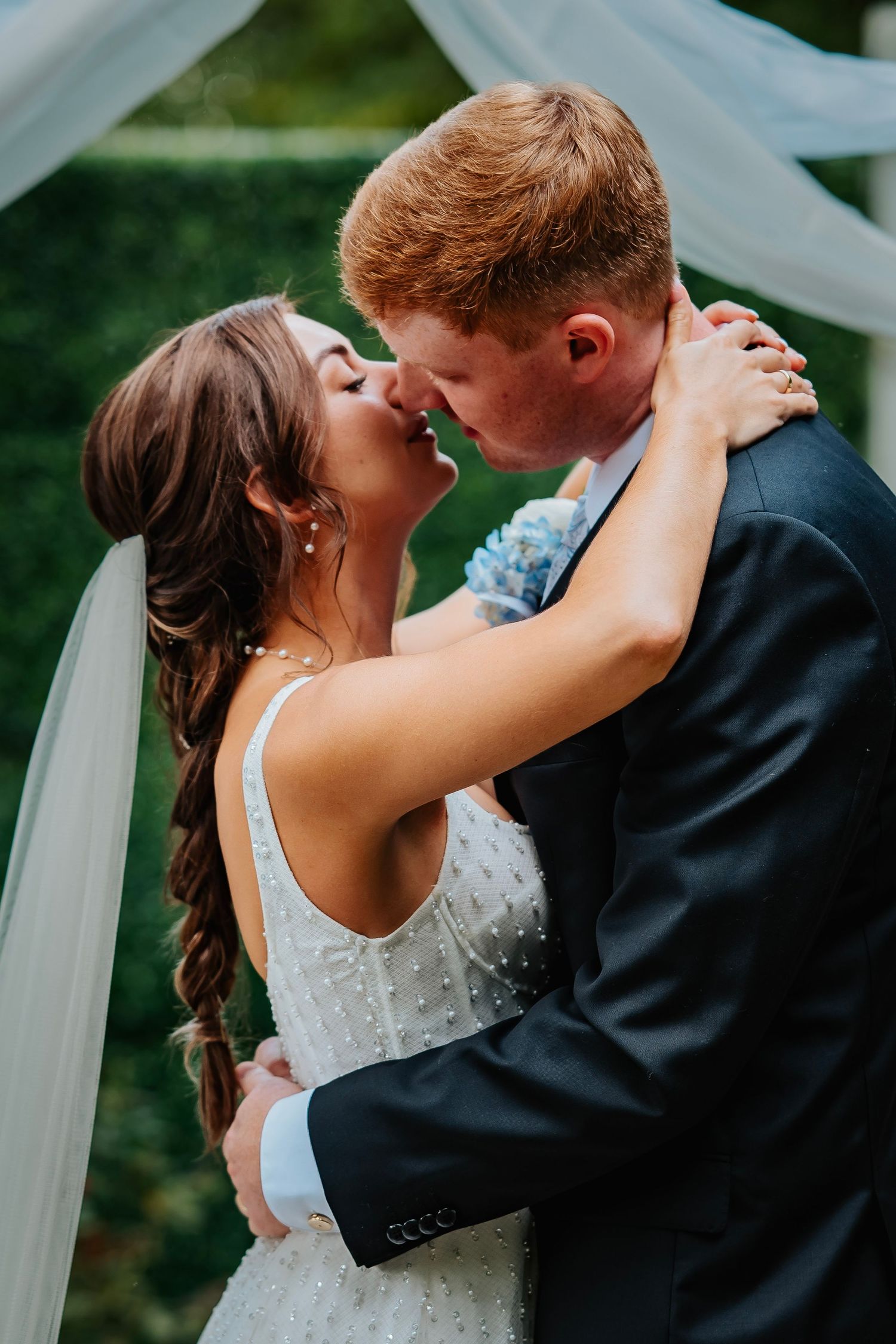 Bride and groom sharing a romantic kiss at their outdoor wedding ceremony under a white draped arch.