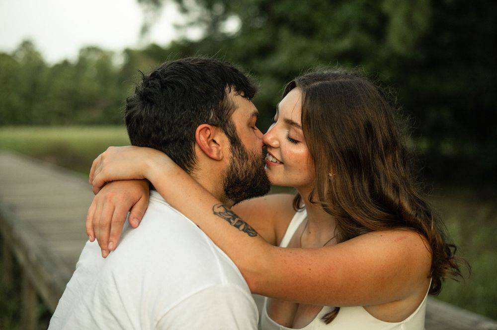 A couple shares a romantic almost-kiss embrace outdoors near a wooden bridge surrounded by lush green trees.