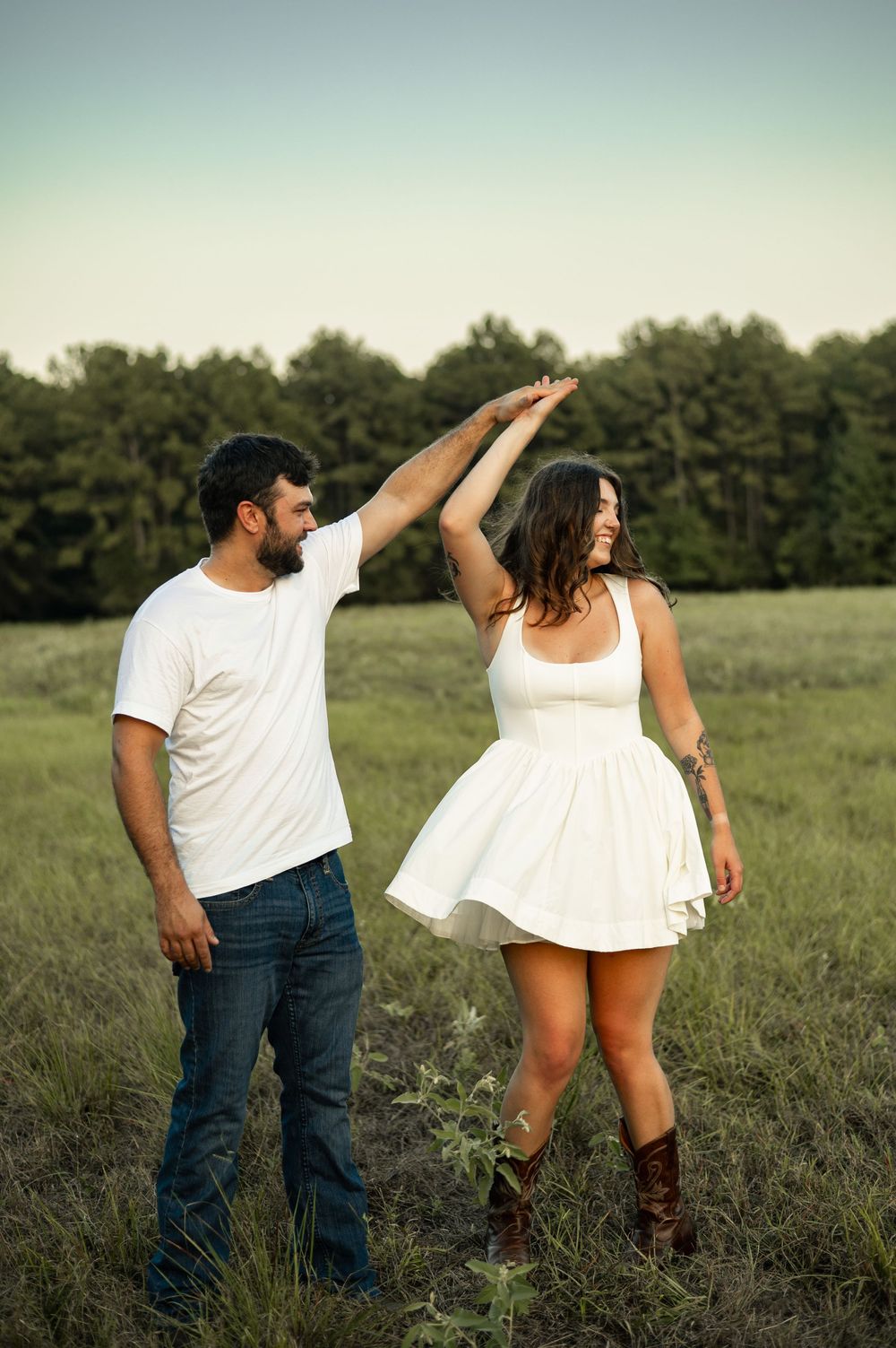 A couple in matching white outfits dance in a green field, man spinning woman in white dress and cowboy boots.