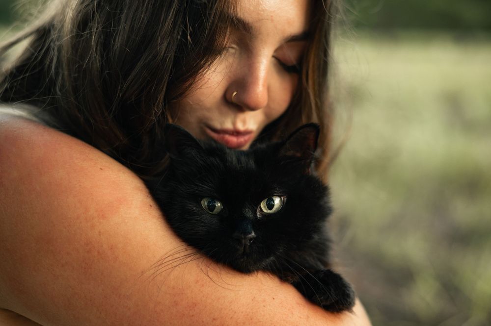 A young woman with long brown hair tenderly holds a fluffy black cat with bright green eyes outdoors.