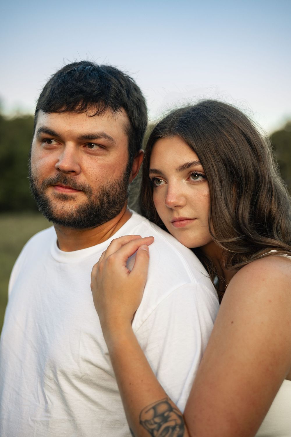 A couple in white shirts pose outdoors at dusk, woman resting hand on man's shoulder with tattoo visible.