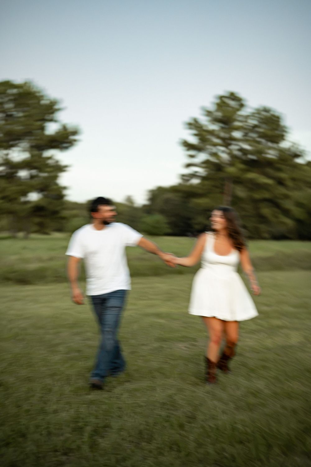 A couple holds hands and walks through a green field with trees in the background on a clear day.