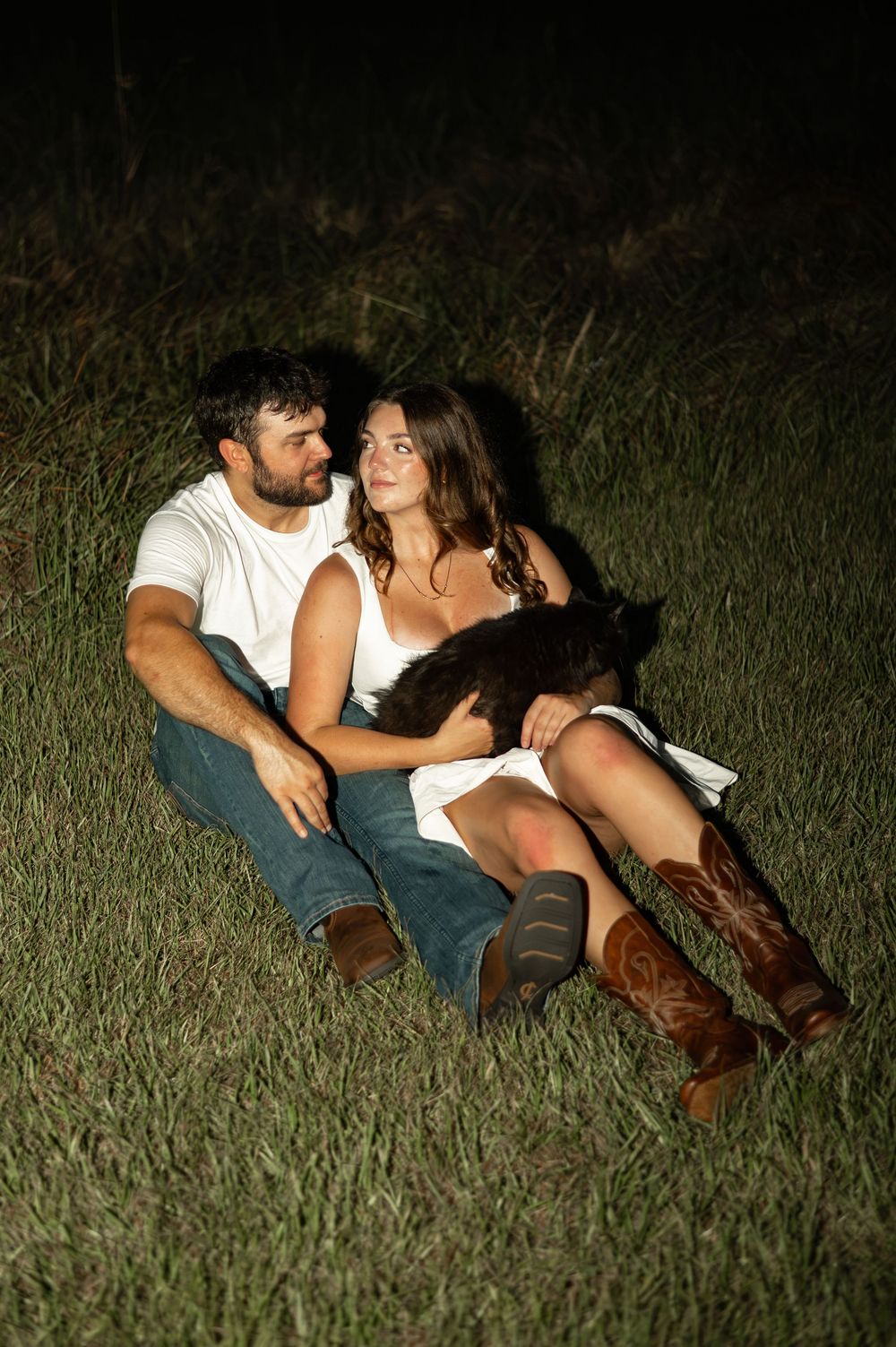 Couple sitting together on grass at night, woman in white dress and cowboy boots holding a black cat, man in white tee.