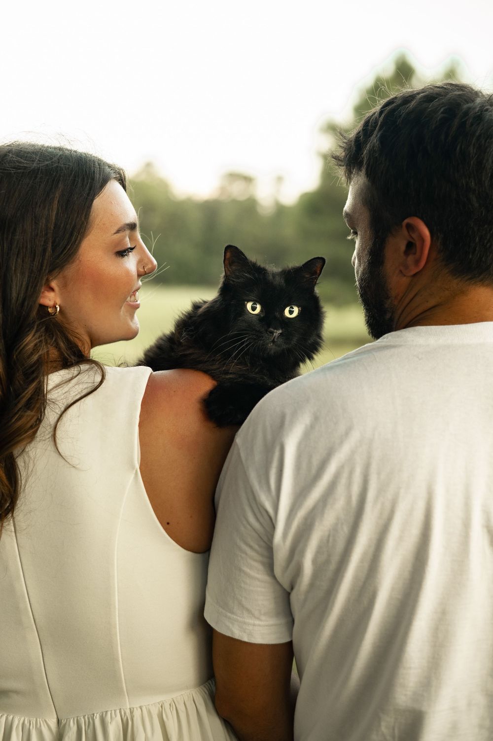 Couple facing each other outdoors holding a fluffy black cat on shoulder, soft natural light background.