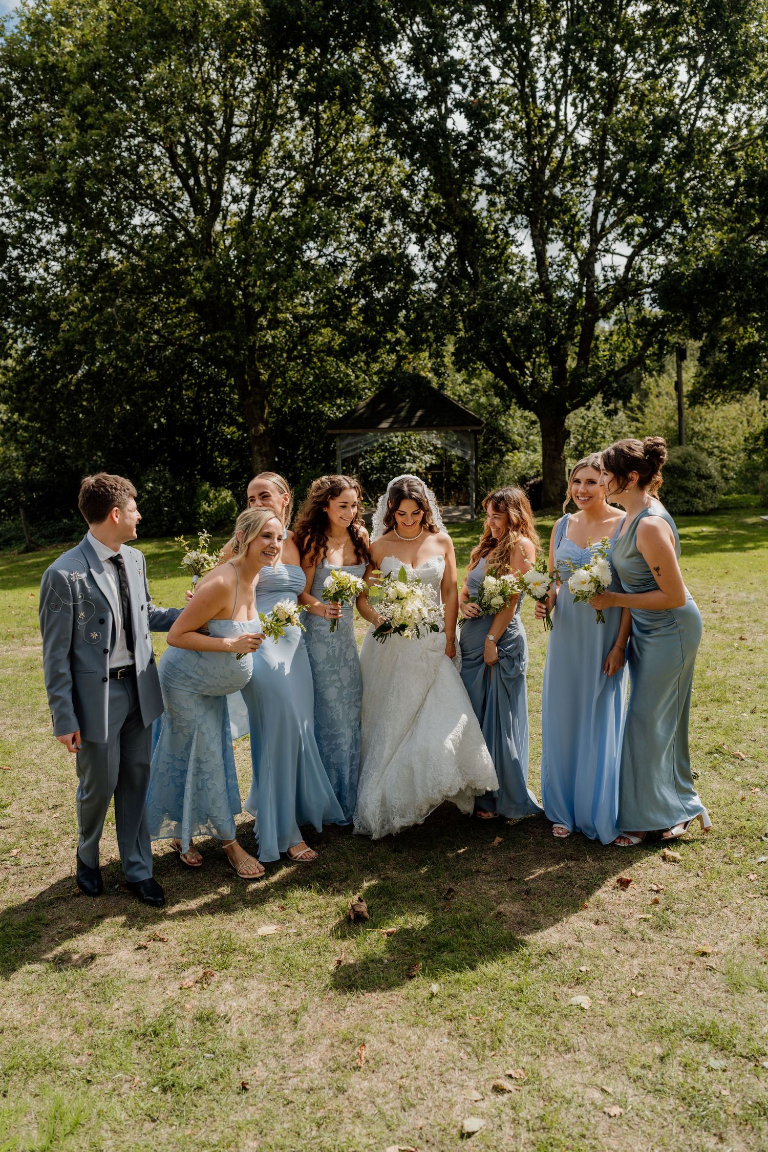 Bride in white gown with bridesmaids in blue dresses holding bouquets outdoors on sunny day.