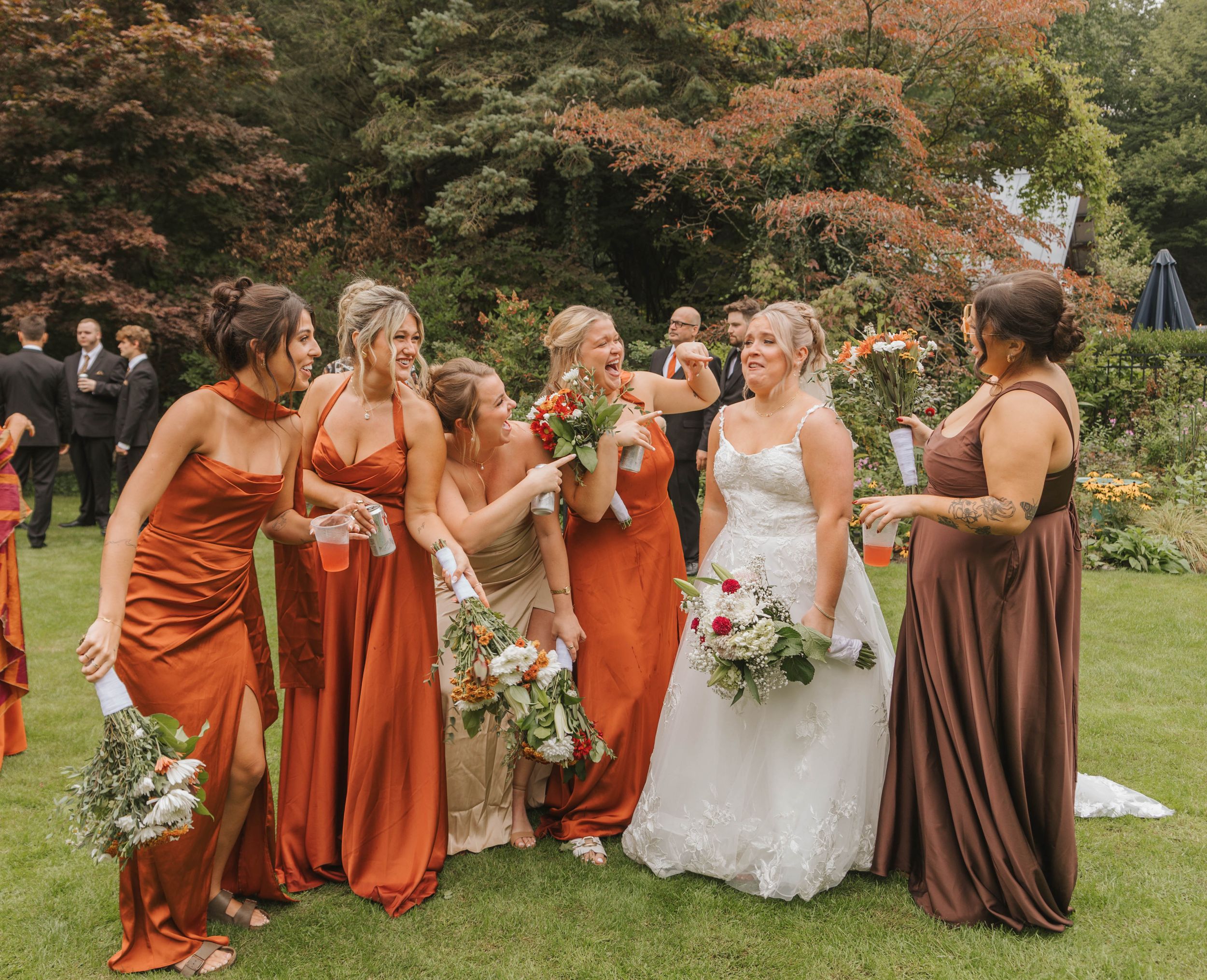 Bride in white gown with bridesmaids in rust and brown satin dresses laughing together on green lawn at outdoor wedding.
