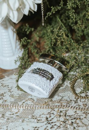 White textured Bella Bianca candle jar with silver lid on lace, surrounded by greenery and white flowers.