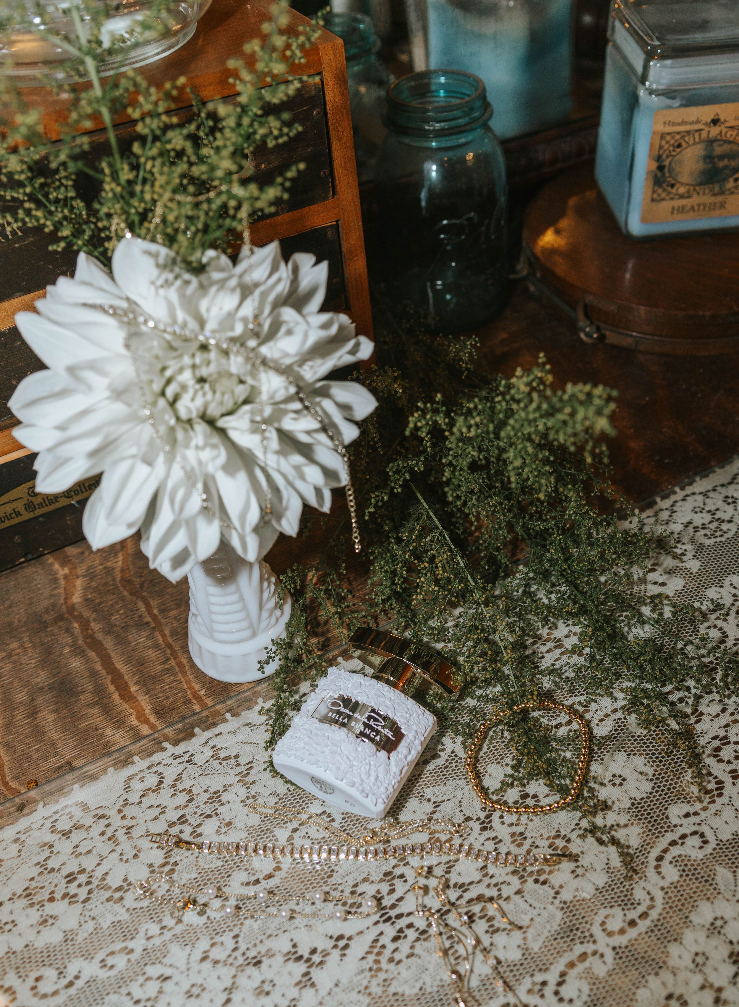 White dahlia in vase with greenery, lace tablecloth, ring box, and gold hoop earrings on rustic wooden table.