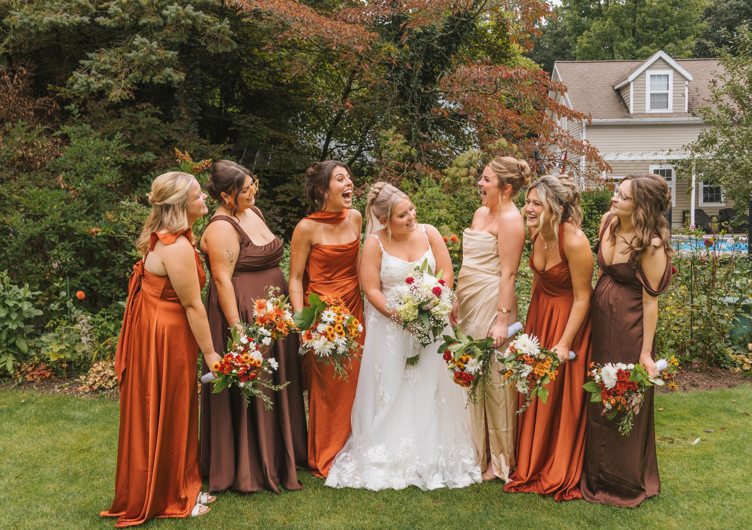 Bride in white gown with bridesmaids in rust and brown dresses holding colorful bouquets in a garden setting.