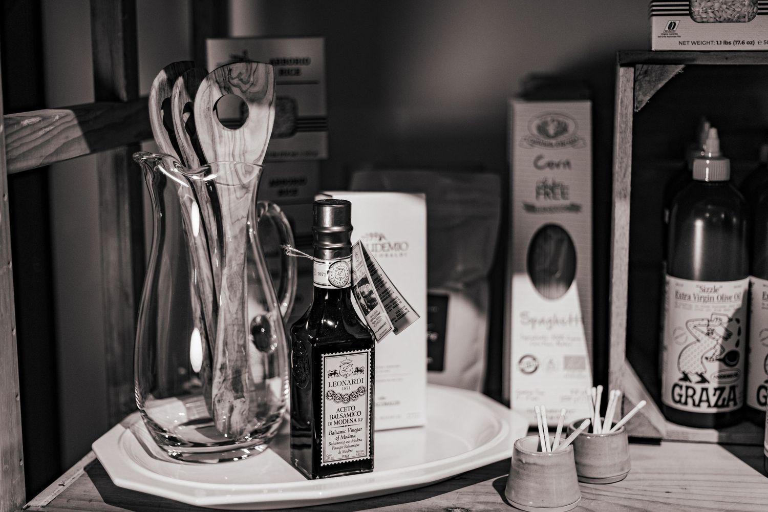 Black and white photo of a kitchen counter with olive oil bottle, utensils in glass vase, and condiments.