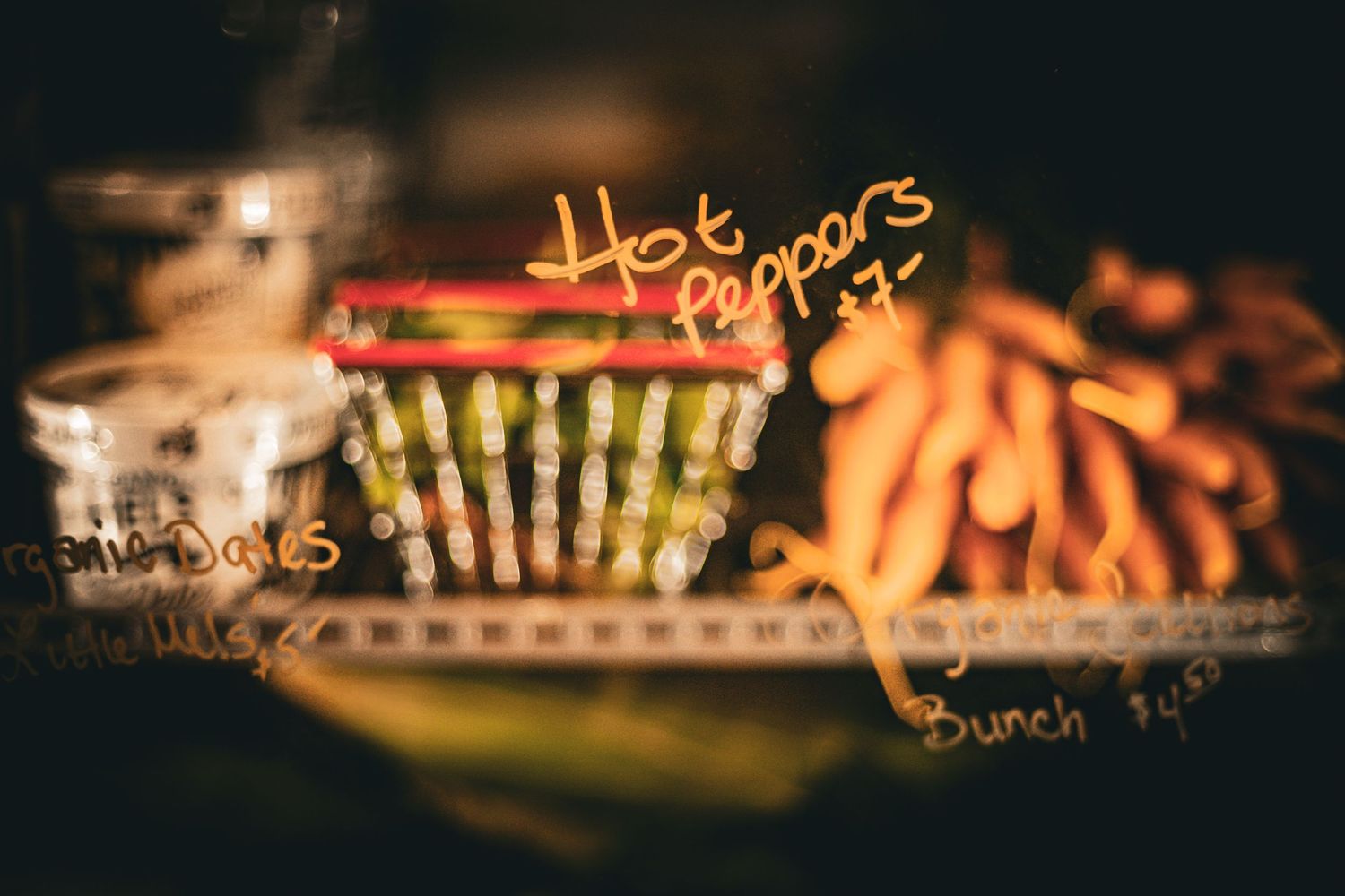 A blurry farmers market display with hot peppers and carrots with handwritten price signs in warm lighting.