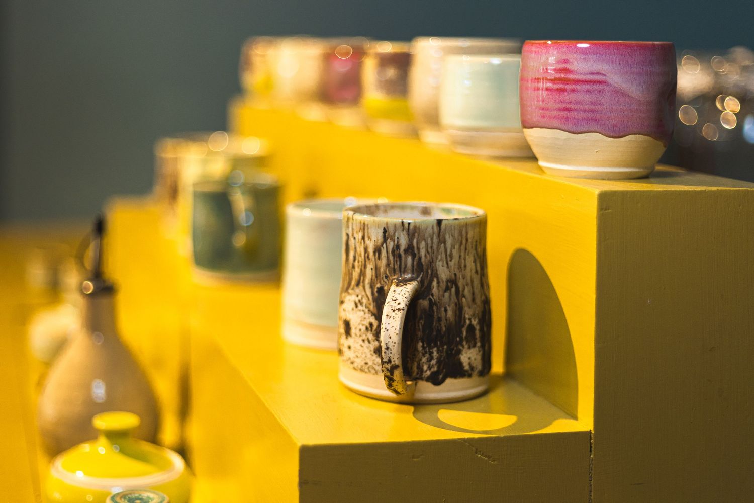 Colorful handmade ceramic mugs and pottery displayed on bright yellow shelves in a modern craft shop.