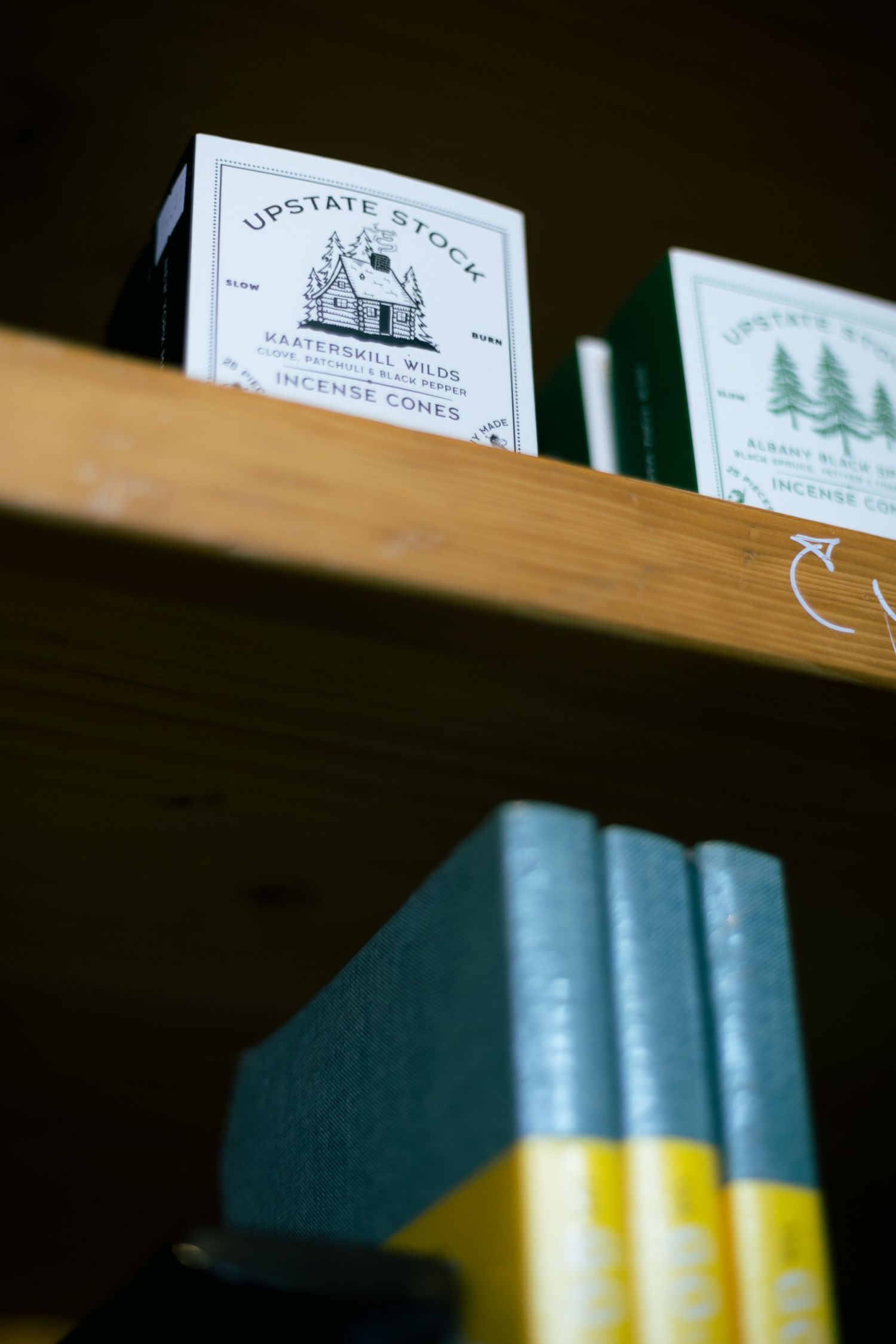 Wooden shelf displaying Upstate Stock incense cone boxes and teal books with yellow spines in a cozy retail setting.