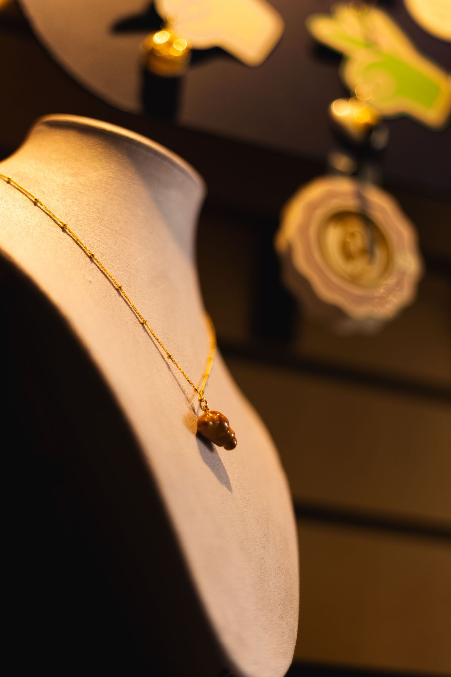Gold chain necklace with amber stone pendant displayed on jewelry bust with blurred accessories in background.