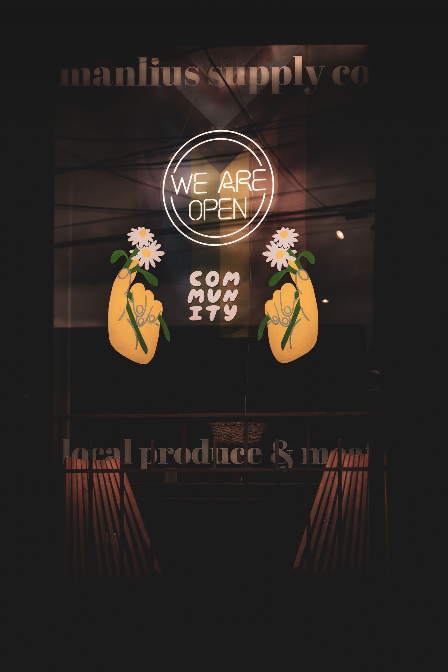 Manlius Supply Co storefront with neon We Are Open sign, community text, and floral hand illustrations on dark window.