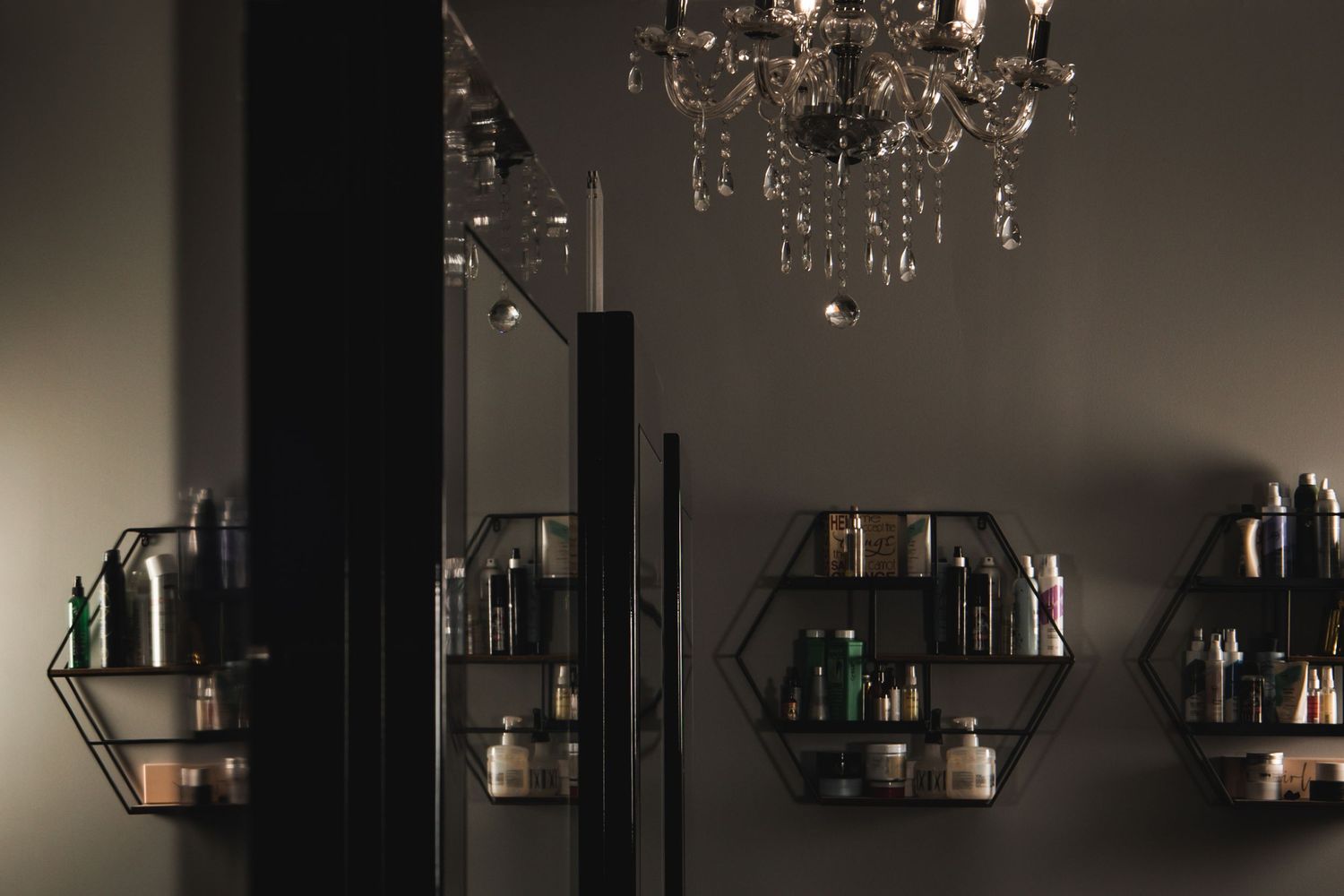 Dark salon interior with hexagonal wall shelves holding beauty products and an elegant crystal chandelier above.