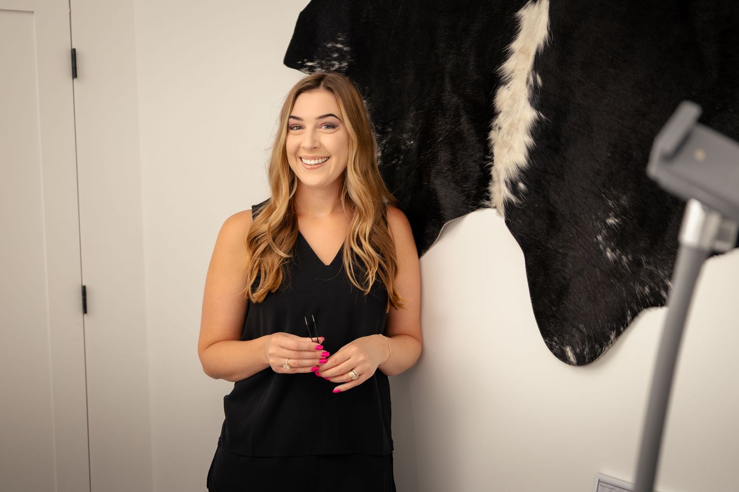 Smiling blonde woman in black sleeveless top holding pink drink, standing near cowhide decor in stylish interior.