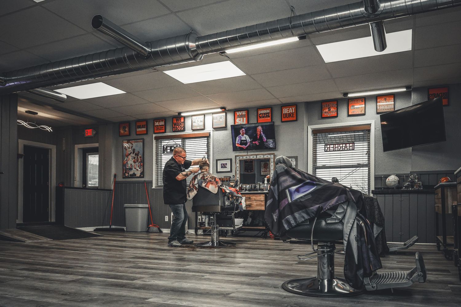 A barber cuts a client's hair in a vintage-style barbershop with framed art, TV, and leather chairs.