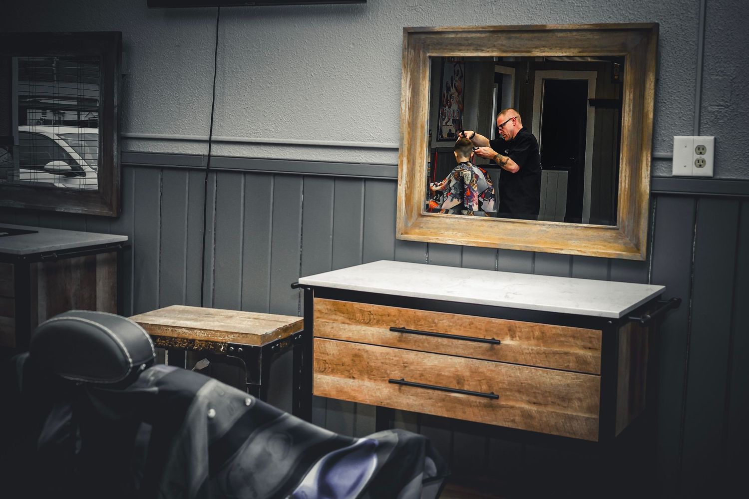 Barber reflected in ornate mirror above rustic wooden dresser in a modern barbershop interior.