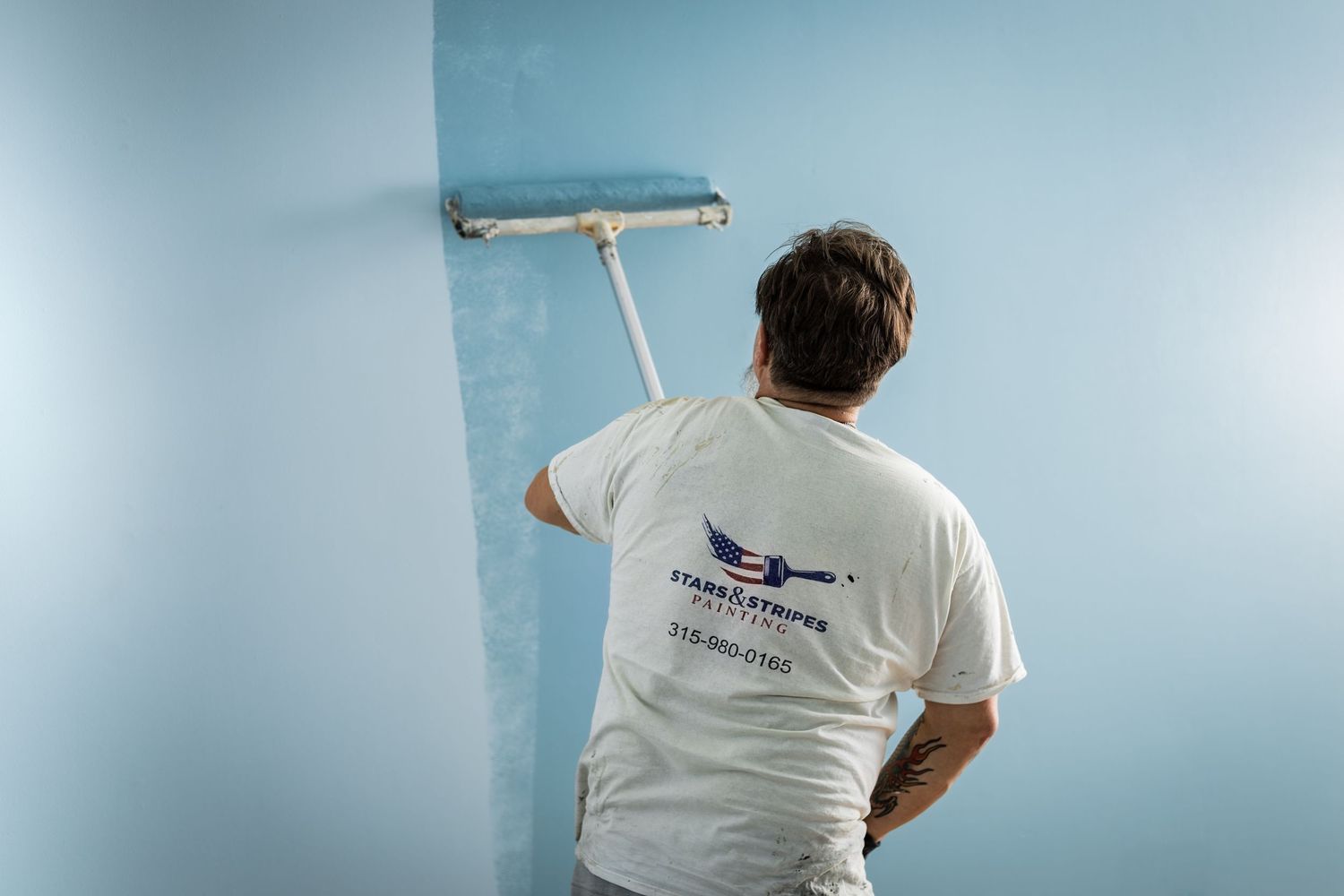 A painter in a white t-shirt uses a roller to apply light blue paint to an interior wall.