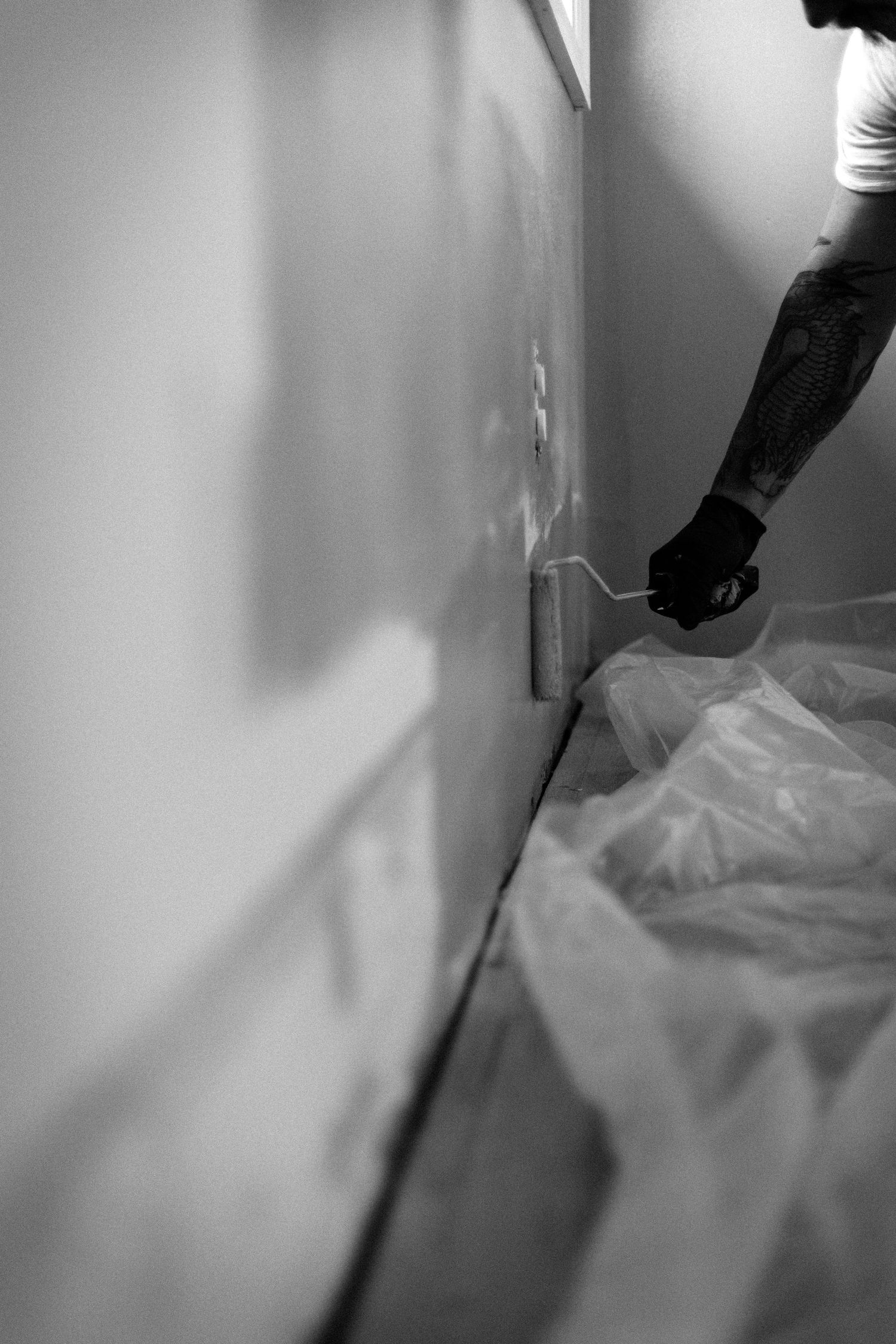 Black and white photo of a tattooed person painting a wall with a roller brush, plastic sheeting on floor.