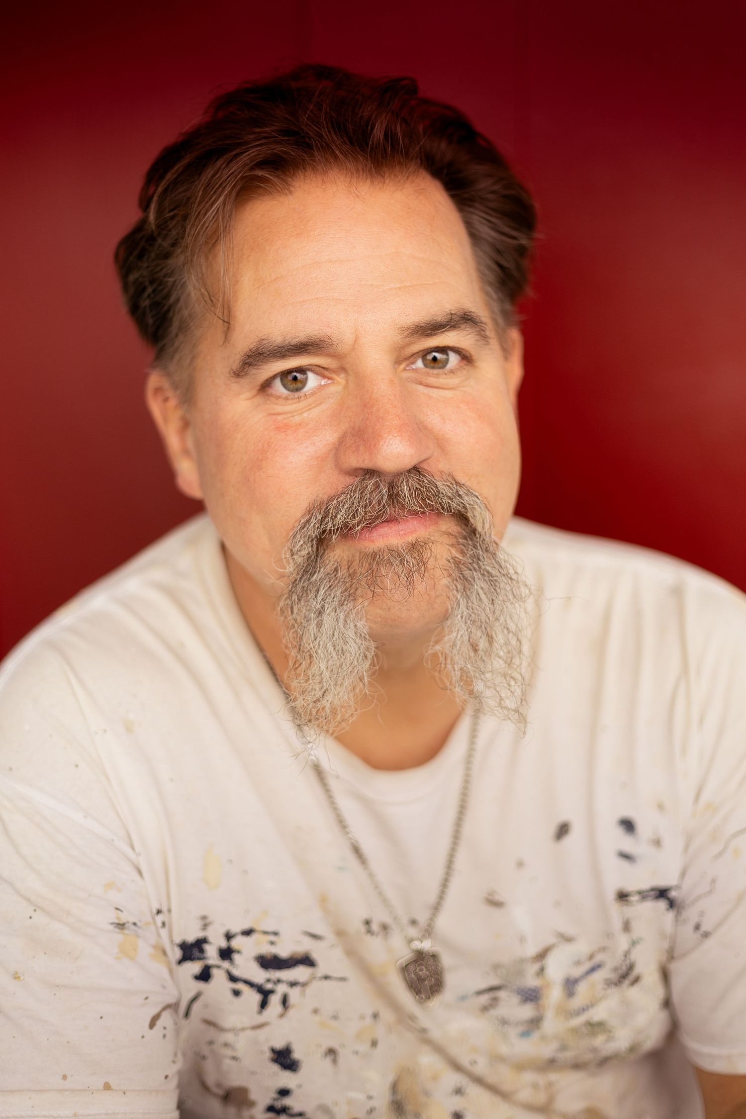 Middle-aged man with gray goatee wearing paint-splattered white t-shirt against red background.