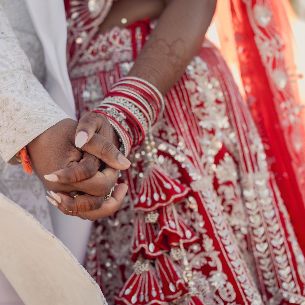 Indian bride and groom holding hands, bride wearing red embroidered lehenga with bangles at traditional wedding ceremony.