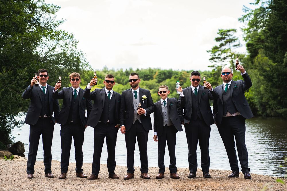 Wedding party in black suits and sunglasses strike casual poses along a scenic waterfront.