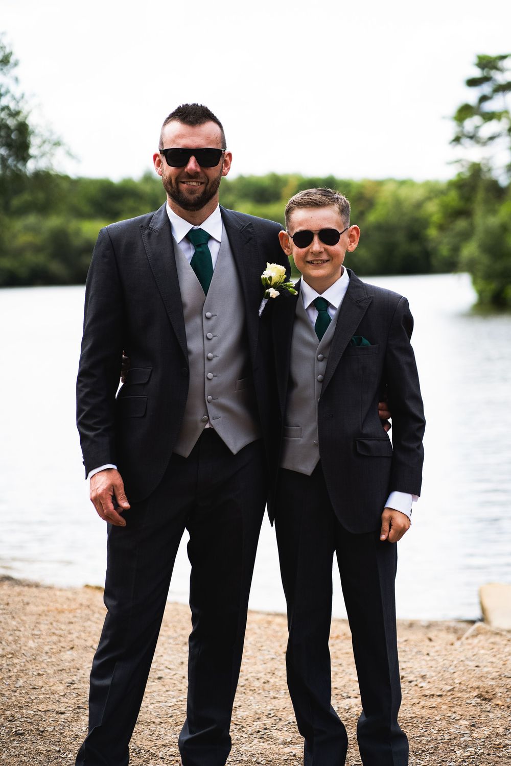 Groomsmen in black suits and sunglasses posing casually by a lakeside beach for wedding party photos.