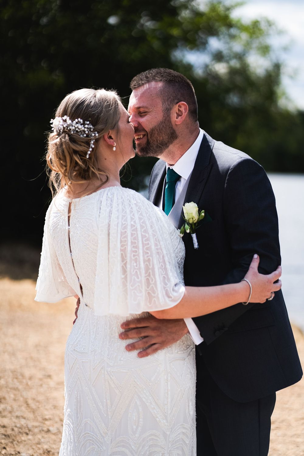 Romantic wedding photo showing a bride in white lace dress with floral hairpiece and groom in navy suit sharing tender moment outdoors.