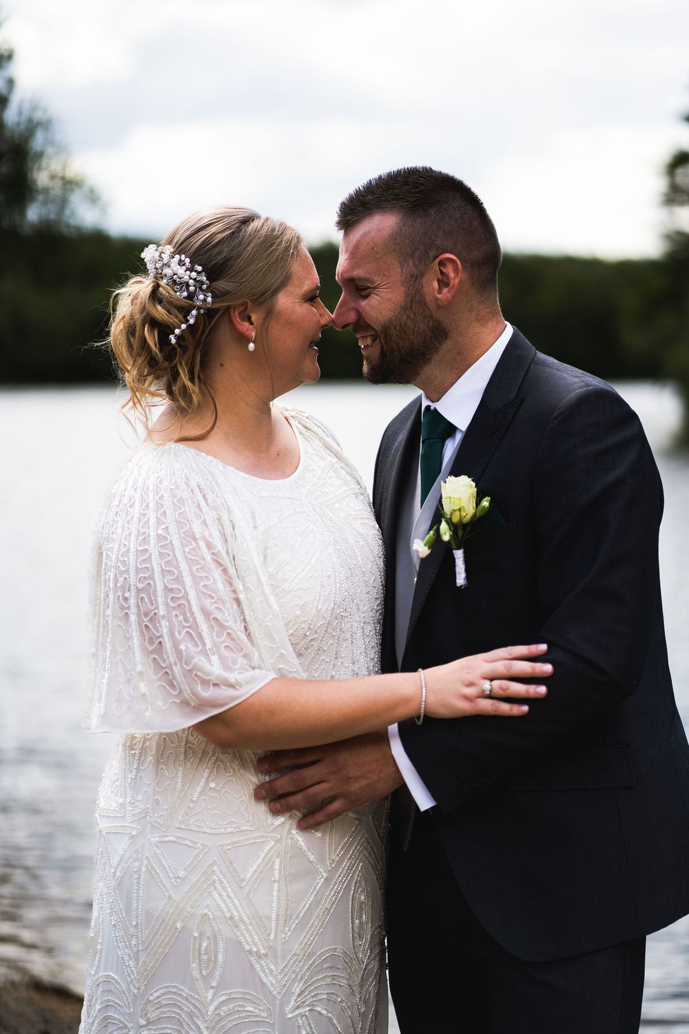Happy newlyweds share an intimate moment by the water, bride in beaded white gown and groom in black suit.