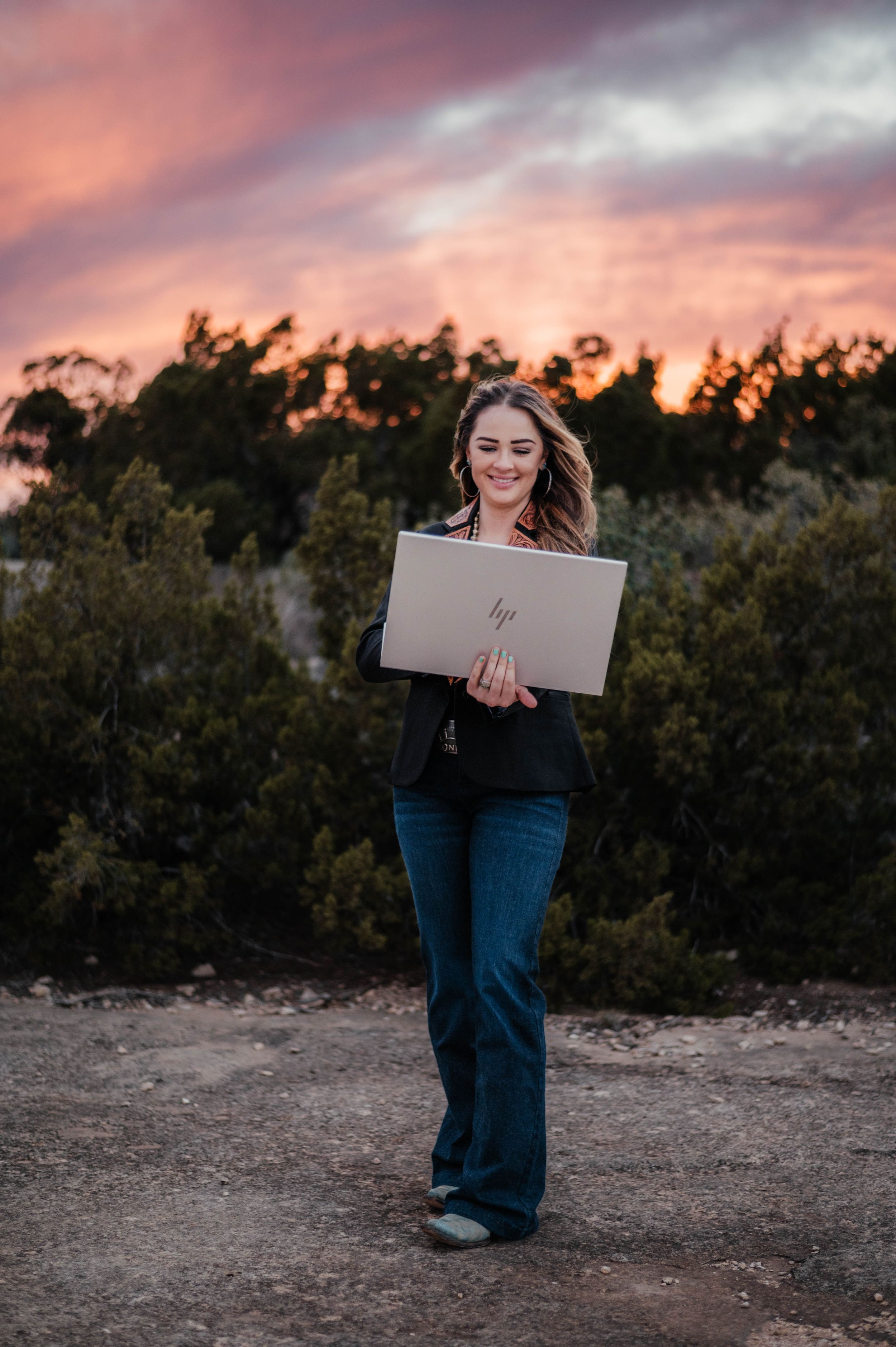 Someone uses a laptop outdoors against a dramatic sunset sky with trees in the background.