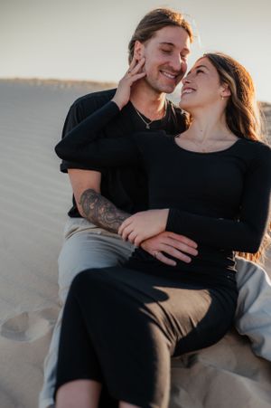 A couple in black clothing sharing an intimate moment while sitting on sandy dunes at sunset.
