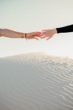 Two hands reaching towards each other against a backdrop of rippling sand dunes in soft lighting.