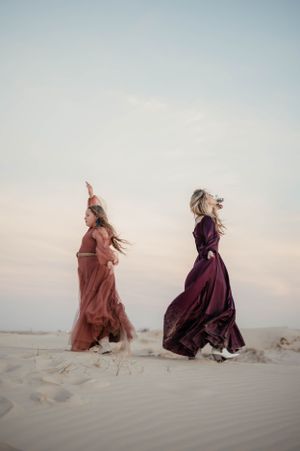 Two women in flowing dresses dancing on sand dunes at dusk.