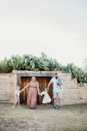 A family stands together in front of a rustic wooden door set in a stone wall with cacti growing above.