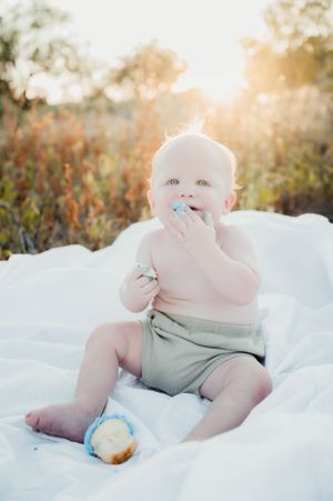 Baby sits on white blanket in soft sunset light with a snack while wearing light-colored shorts and eating.
