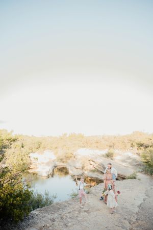 Silhouetted figures walk along a rocky cliff path overlooking a tranquil pond under a soft, hazy sky.