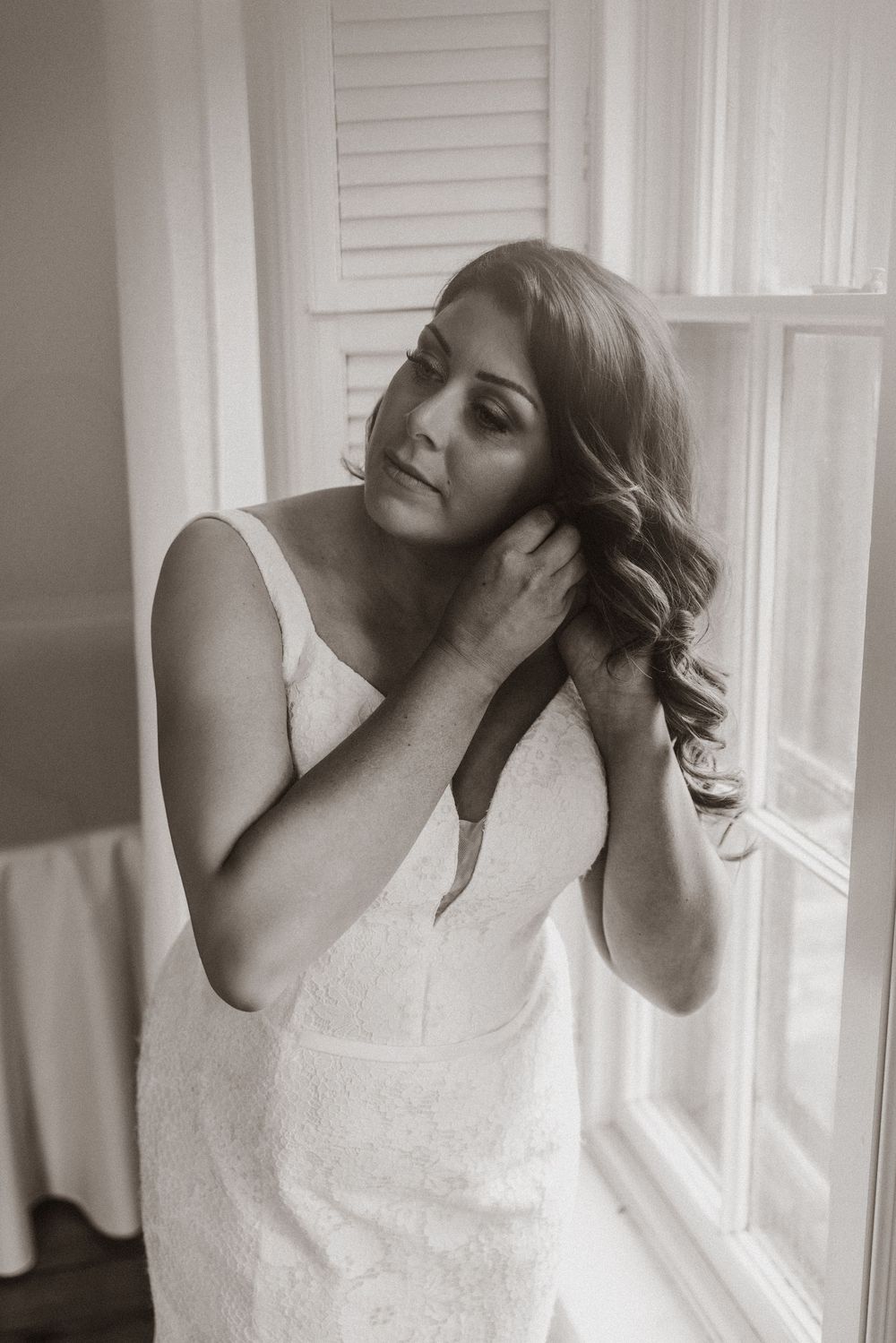 Bride in white dress poses gracefully by window in soft natural light.