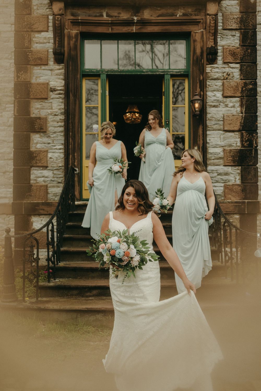 Bridal party in flowing dresses stands outside stone mansion entrance.